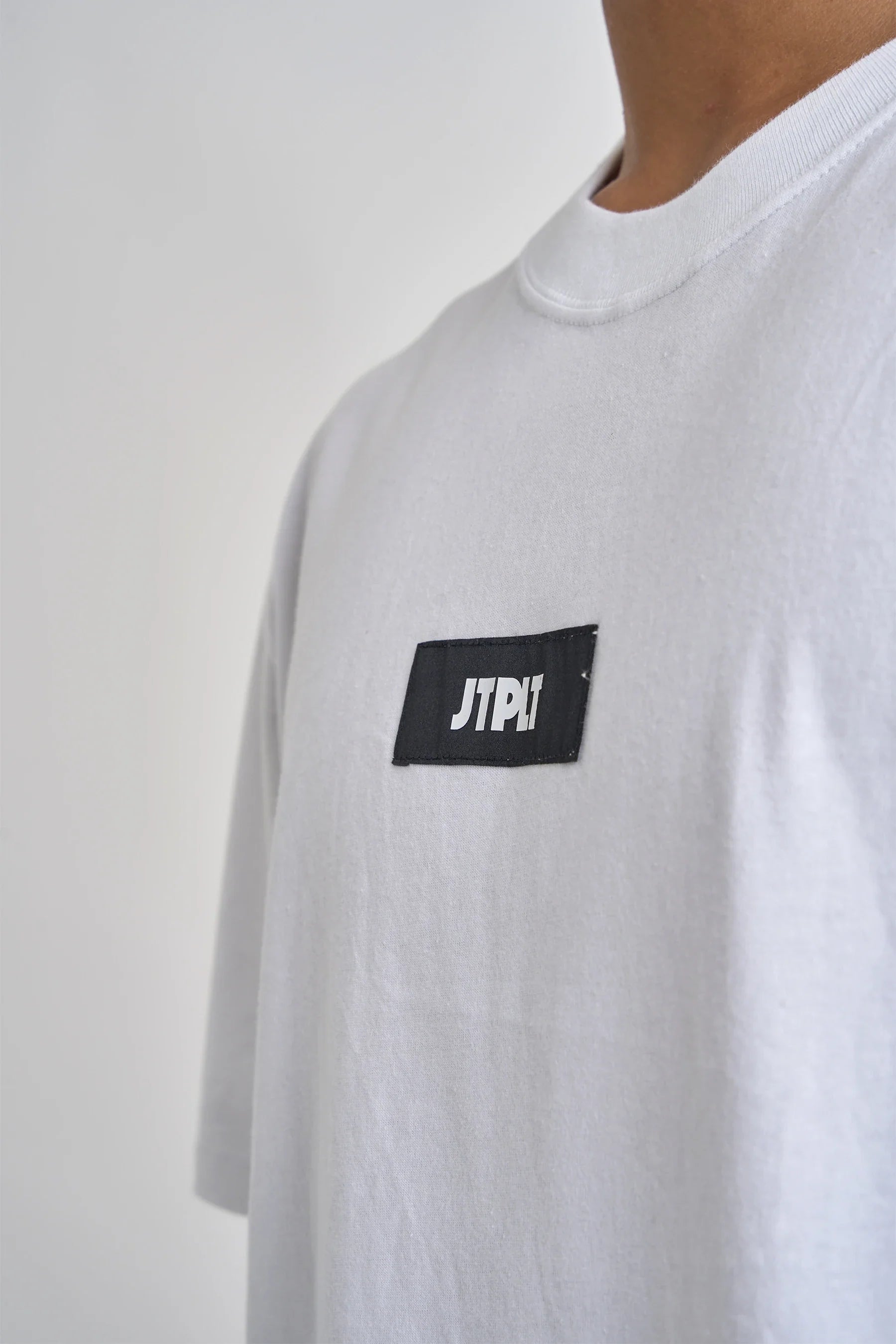 White t-shirt with a black patch featuring 'JTPLT' on a plain background