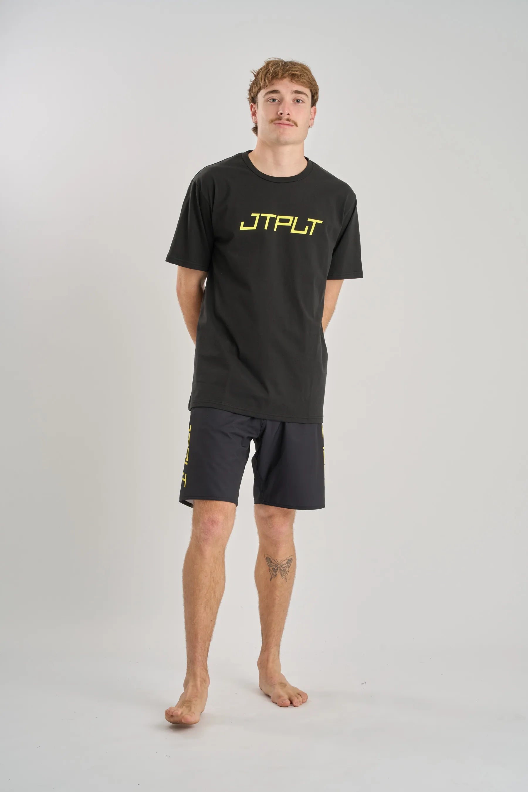 Man wearing a black t-shirt with 'JTPLT' logo and black shorts on a grey background