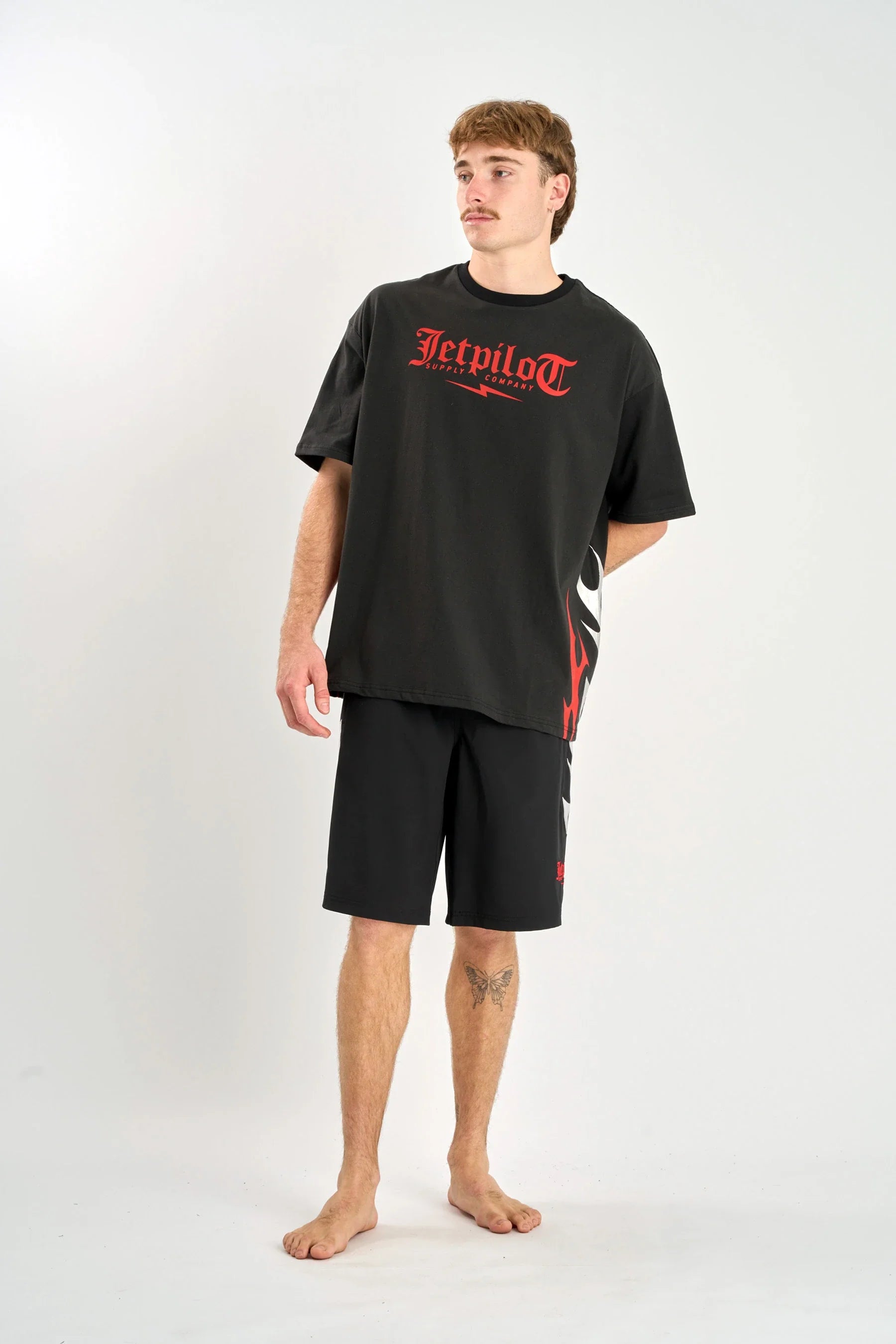 Person wearing a Jetpilot Tribal Heavy Tee - Black with red text and black shorts on a white background
