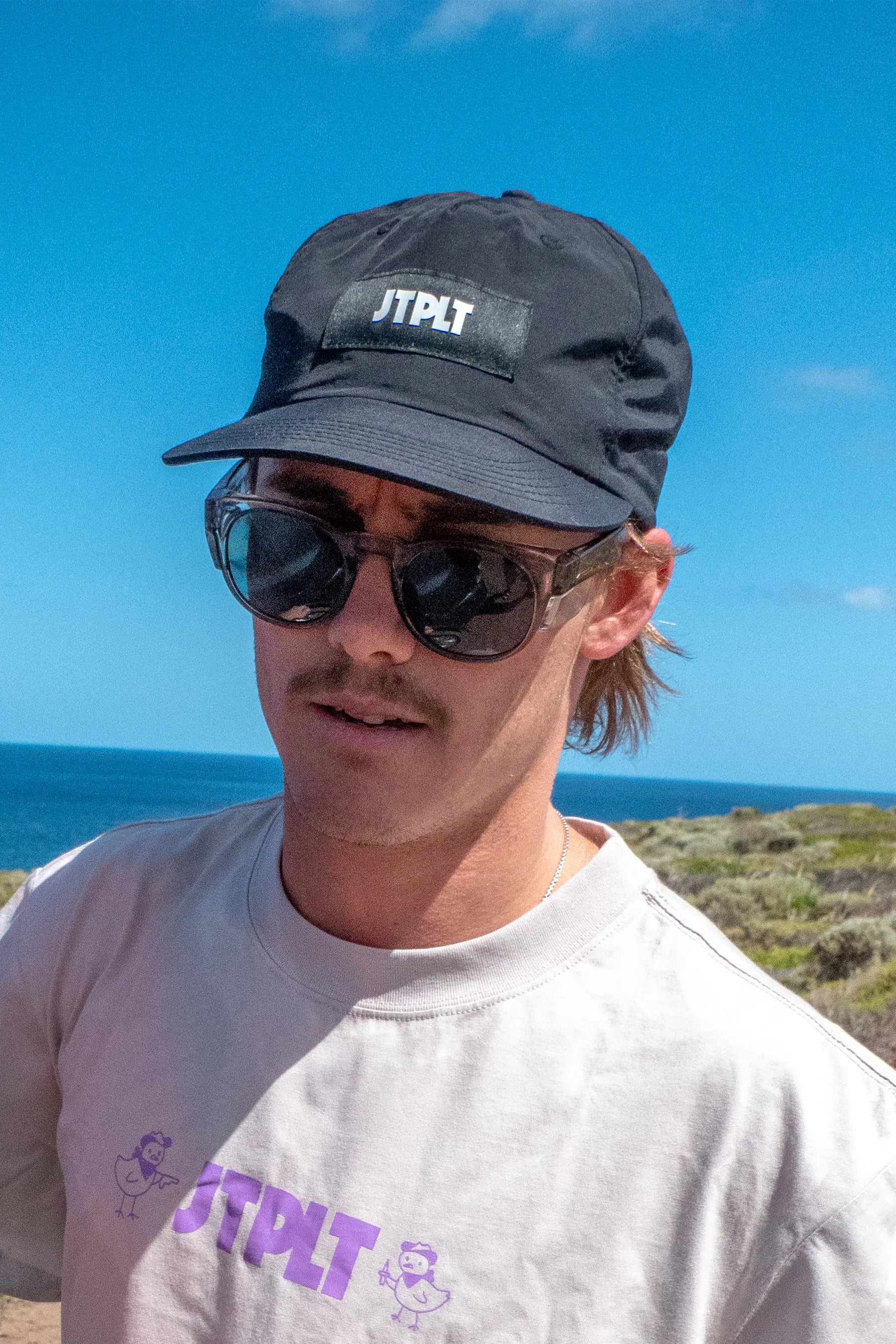 Person wearing a black JTPLT branded cap and sunglasses with a blue sky and ocean background