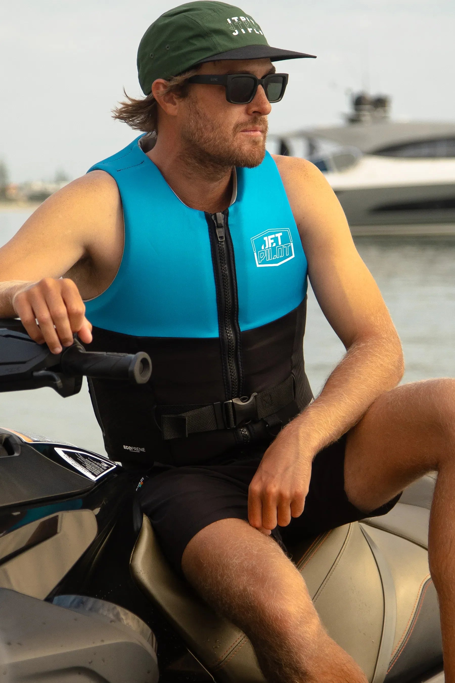 Man wearing a blue life jacket and green cap sitting on a jet ski by the water.