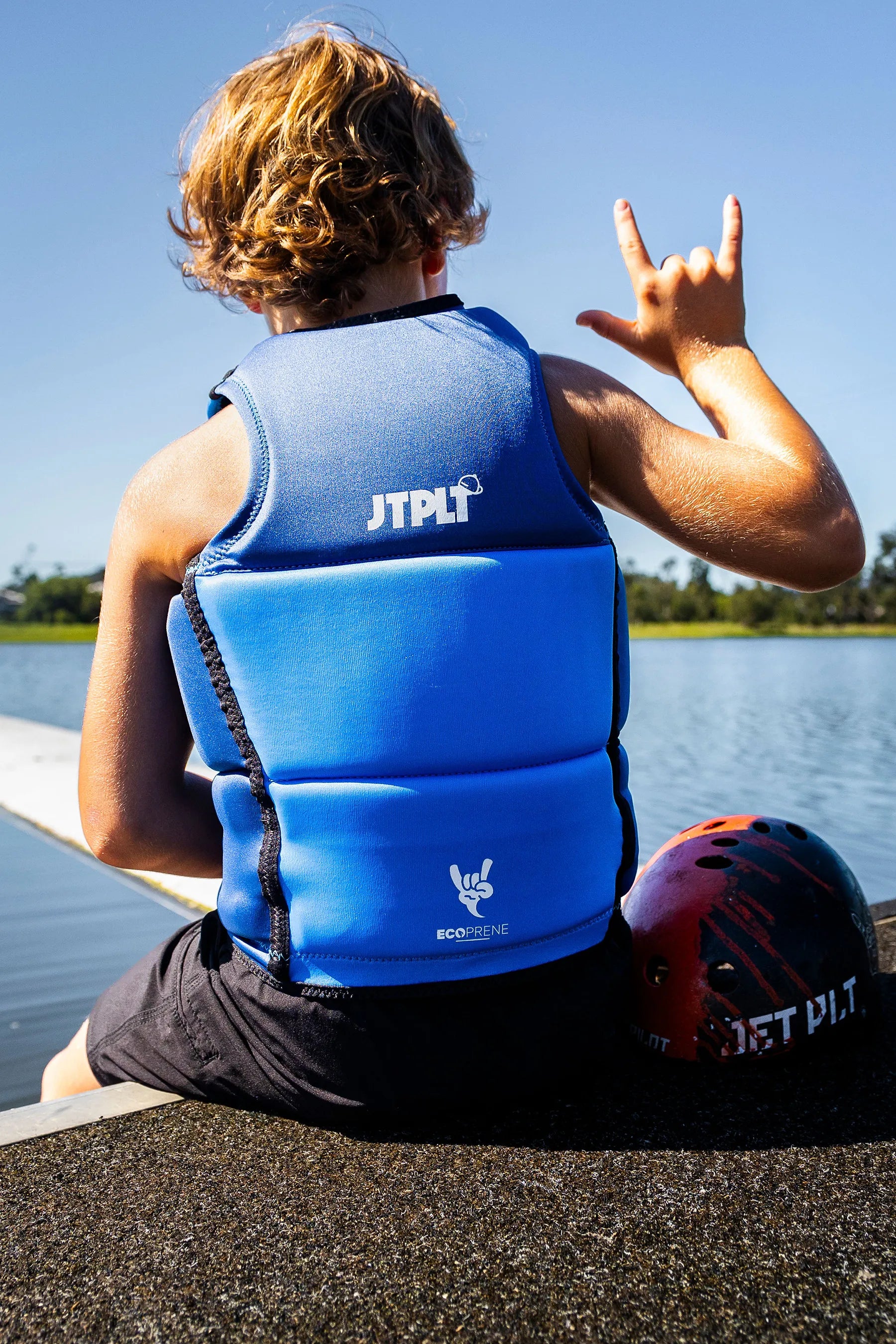 Child wearing a Jetpilot Freeride Boys Life Jacket - Blue by a lake, making a rock-on sign.
