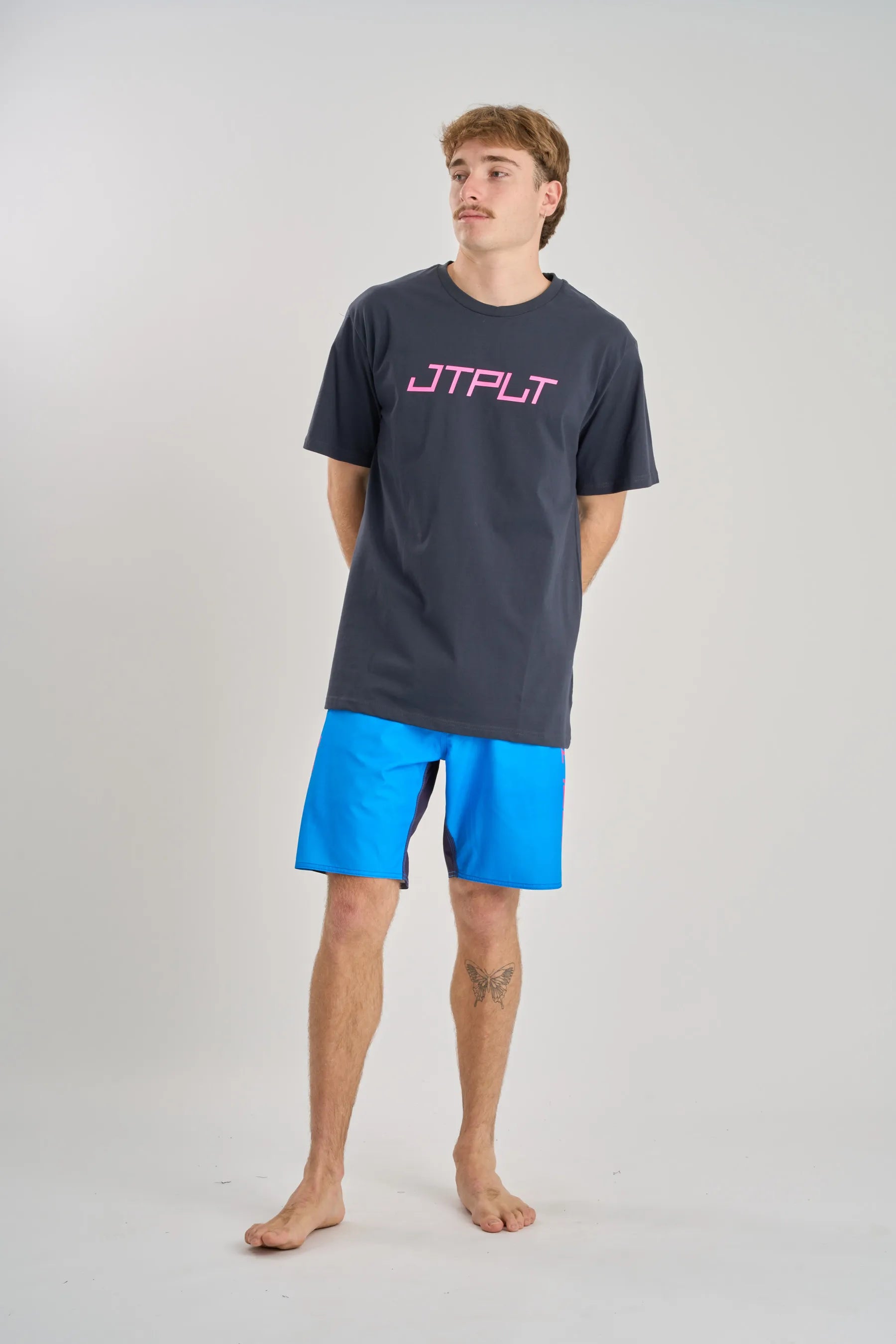 Man wearing a dark gray t-shirt with 'JTPLT' logo and blue shorts on a white background