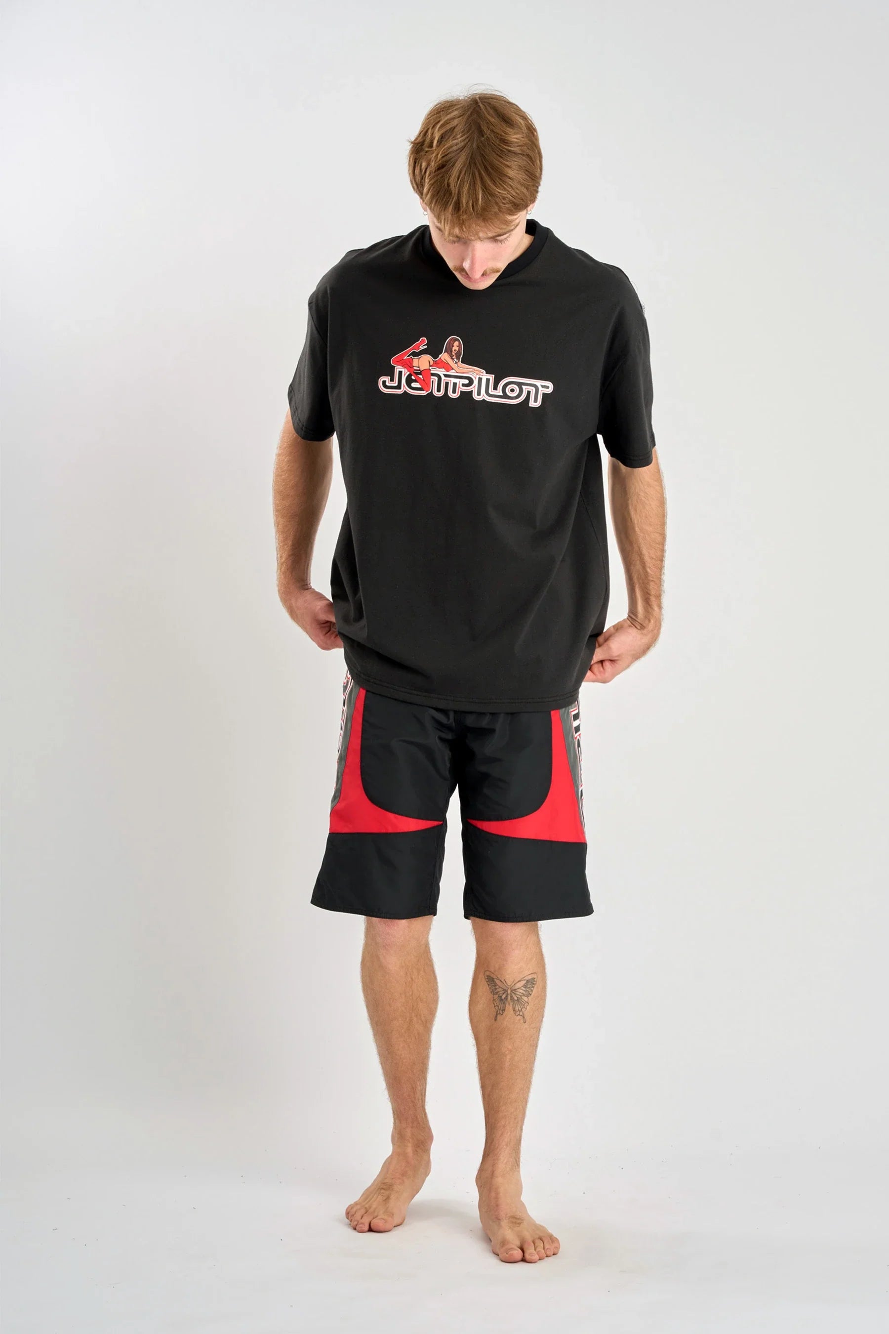 Person wearing a Jetpilot Moto Heavy Tee - Black with a logo and red shorts on a white background