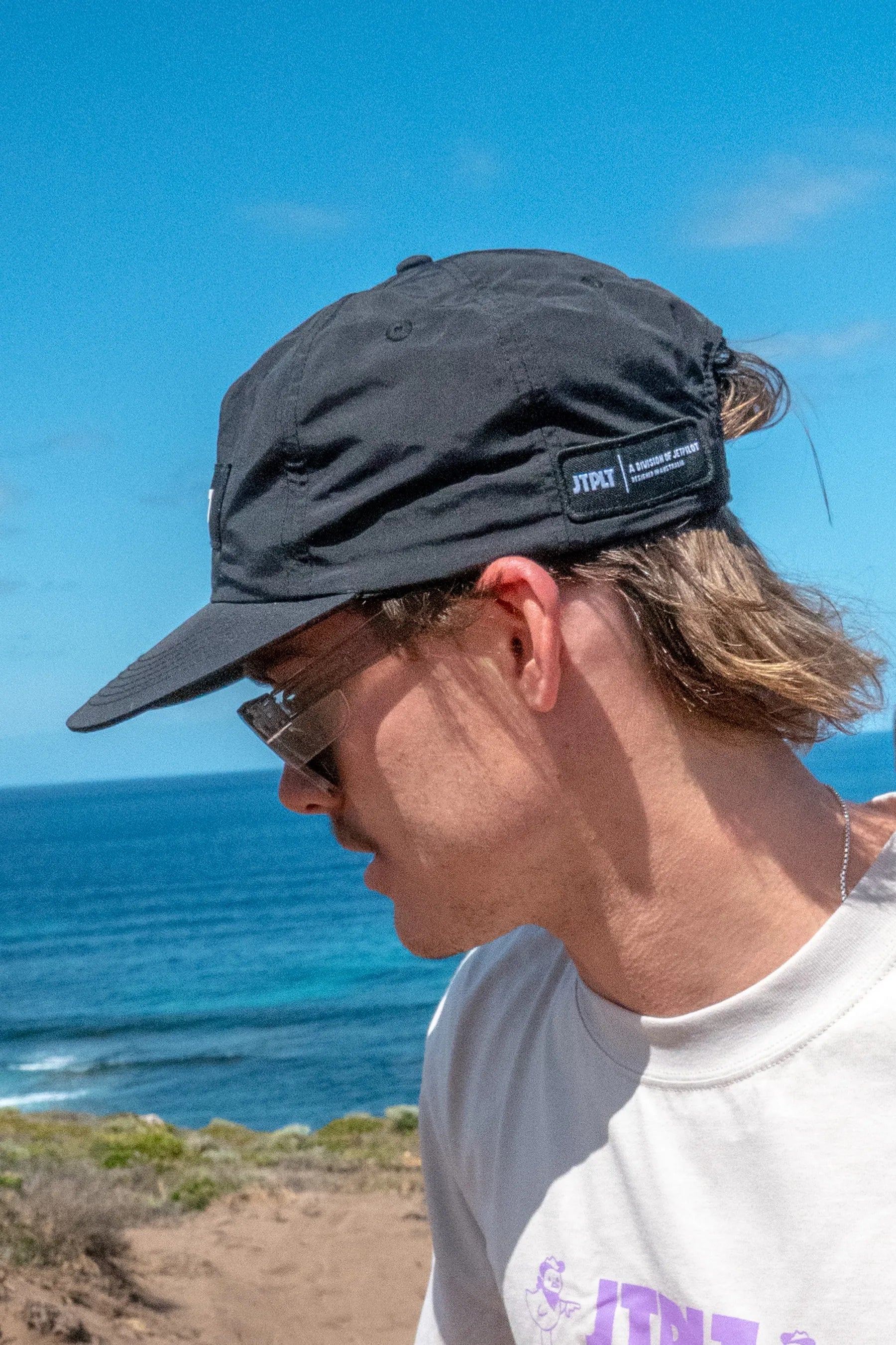 Person wearing a black cap with a logo, standing on a coastal path with blue sky and ocean.