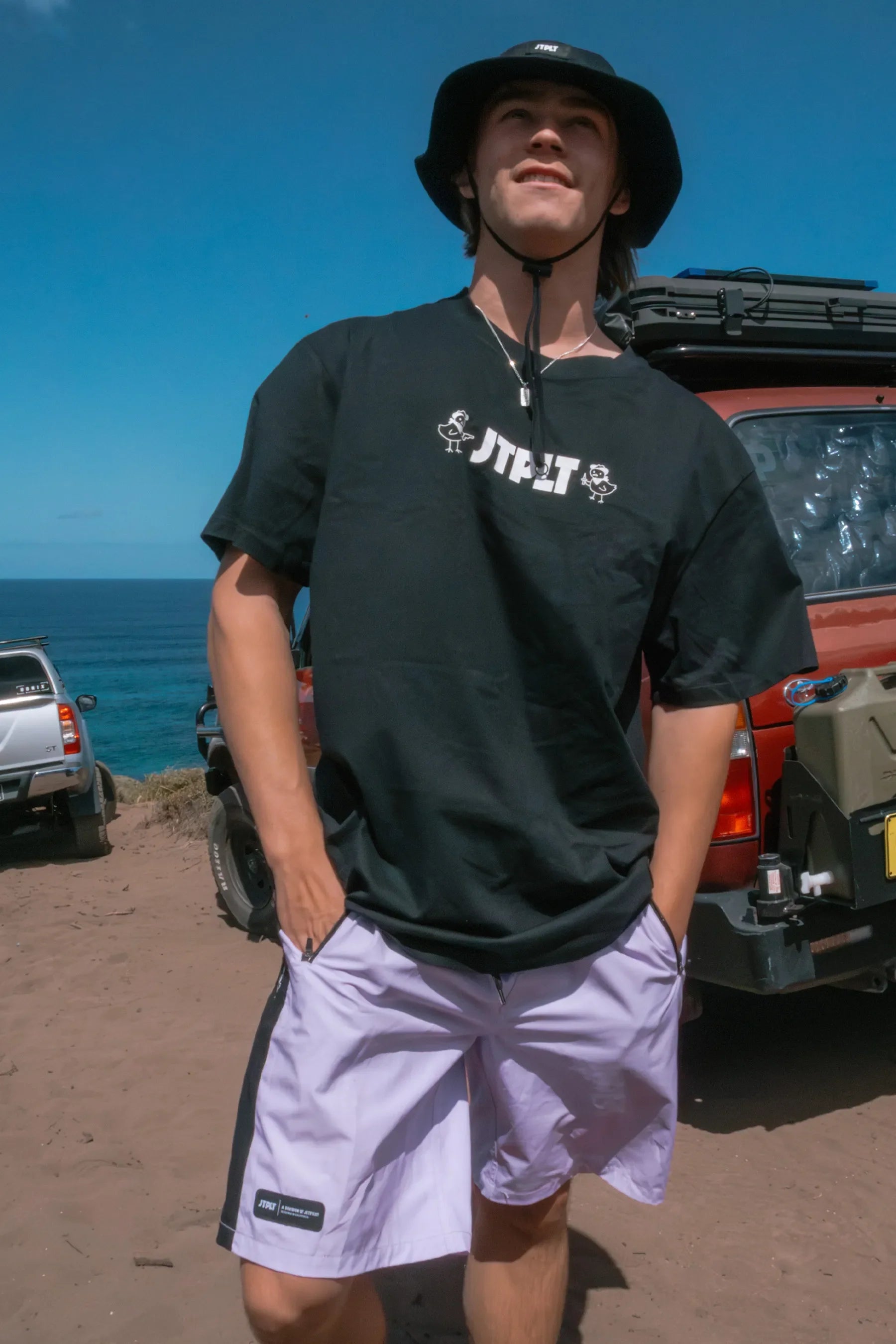 Person wearing a Jetpilot Chicken Tee - Black t-shirt and purple shorts standing in front of a vehicle with a scenic background.