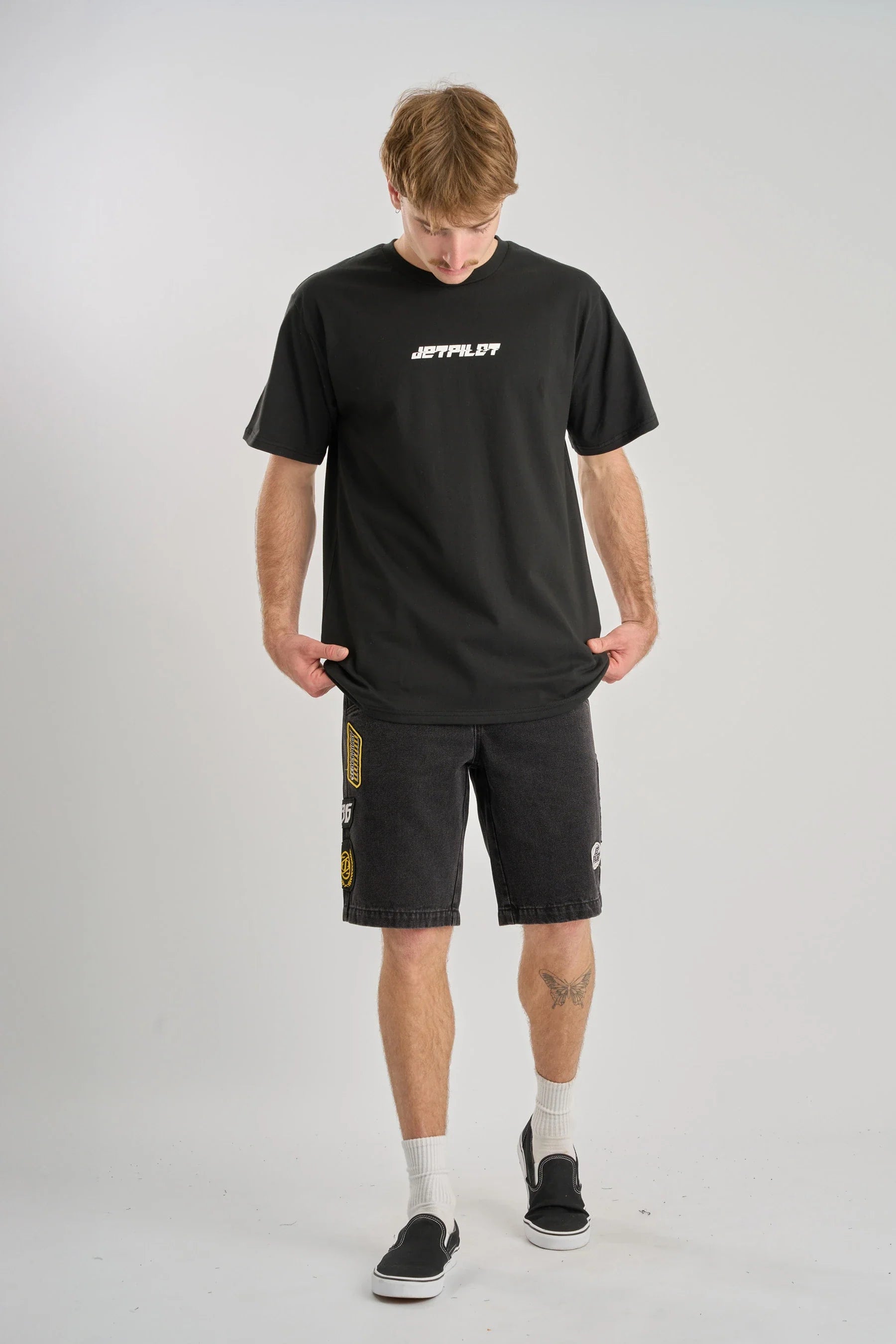 Man wearing a black Jetpilot Progressive t-shirt and shorts with a logo on a grey background